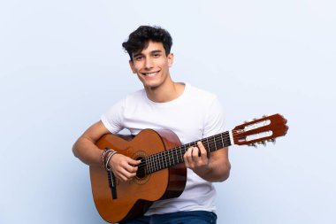 İzole edilmiş mavi arka planda gitar çalan genç Arjantinli adam çok gülümsüyor.