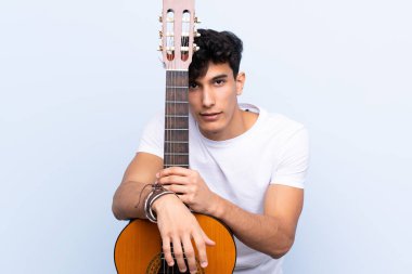 İzole edilmiş mavi arka planda gitar çalan genç Arjantinli adam.