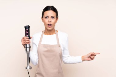 İzole edilmiş arkaplanda el blenderi kullanan kadın şaşırmış ve işaret ediyor.