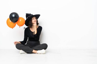 Elinde siyah ve turuncu hava balonları olan genç cadı yere oturmuş gülümserken yukarı bakıyor.