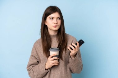 İzole edilmiş mavi arka plan üzerine kazak giyen genç Ukraynalı kız elinde kahve ve bir şeyler düşünürken bir cep telefonu tutuyor.