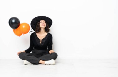 Genç cadı elinde siyah ve turuncu hava balonları yerde oturmuş gülüyor.