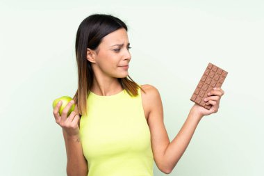 Bir elinde çikolata tableti, diğer elinde elma tutarken yeşil arka planda kalan kadının şüpheleri var.