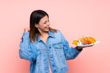 İzole edilmiş pembe arka planda waffle tutan esmer kadın zaferi kutluyor.
