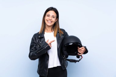 Genç esmer kadın, izole edilmiş mavi arka planda motosiklet kaskı tutuyor ve ona doğrultuyor.