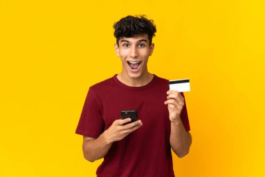 Arjantinli genç adam sarı arka planda izole edilmiş cep telefonuyla alışveriş yapıyor ve elinde şaşırmış bir ifadeyle bir kredi kartı tutuyor.
