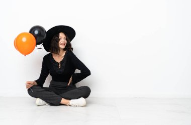 Siyah ve turuncu hava balonları tutan genç cadı kolları kalçasında poz veriyor ve gülümsüyor.