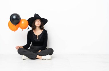 Elinde siyah ve turuncu hava balonları olan genç cadı yerde oturuyor ve yukarı bakıyor.