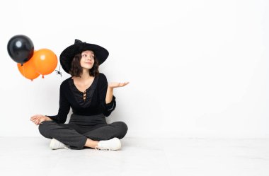 Siyah ve turuncu hava balonları tutan genç cadı yerde oturuyor ve ellerini kaldırırken kuşkuları var.