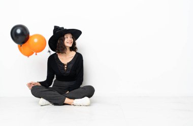 Siyah ve turuncu hava balonları tutan genç cadı yere oturmuş yan tarafa bakıyor ve gülümsüyor.