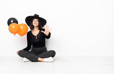 Yerde oturan siyah turuncu balonları tutan genç cadı parmaklarıyla beşi sayıyor.