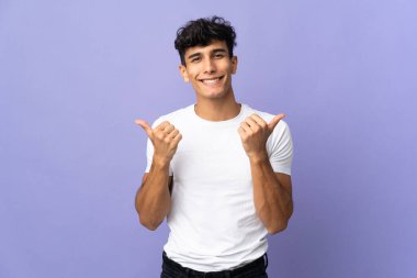Arjantinli genç adam arka planda izole edilmiş. Baş parmakları havada ve gülümsüyor.