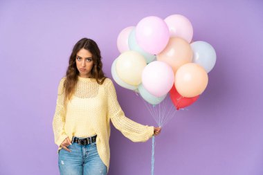 Mor arka planda izole edilmiş bir sürü balon yakalayan genç kadın üzgün.