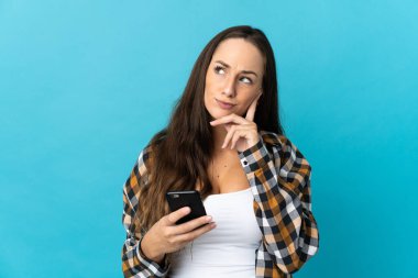 İzole edilmiş mavi arka plan üzerinde cep telefonu kullanan ve düşünen genç İspanyol kadın.