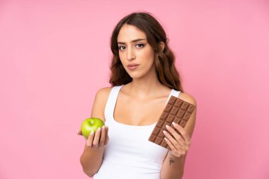 Soyutlanmış pembe arka planda genç bir kadın bir elinde çikolata tableti, diğerinde elma alıyor.