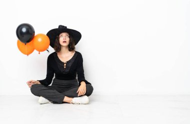 Elinde siyah ve turuncu hava balonları olan genç cadı yere oturmuş ve şaşırmış bir ifadeyle bakıyor.