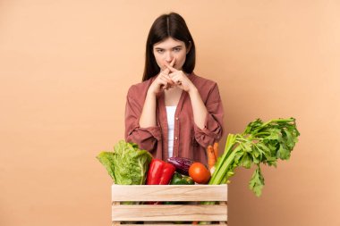 Bir kutuda taze sebze toplayan genç çiftçi kız sessizlik işareti gösteriyor.