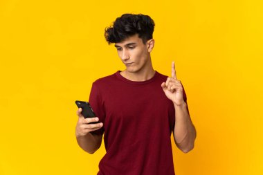 Arjantinli genç adam sarı arka planda cep telefonu ve parmak kaldırarak izole edildi.