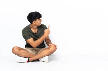 Genç Arjantinli bir adam yerde oturmuş işaret parmağıyla işaret ediyor.