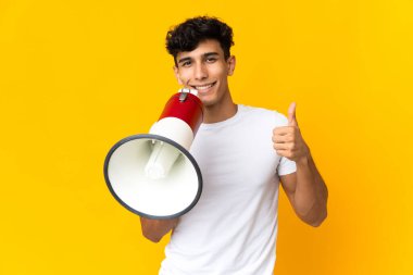 Sarı arka planda izole edilmiş genç Arjantinli bir adam baş parmağıyla bir megafon tutuyor.