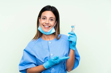 İzole edilmiş yeşil arka planda aletleri tutan dişçi kadın davet edilmek için ellerini yana uzatıyor.