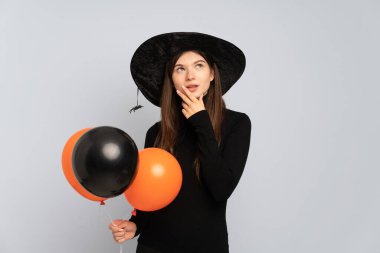 Siyah ve turuncu hava balonları tutan genç cadı gülümserken yukarı bakıyor.