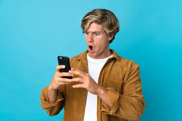 English man over isolated blue background looking at the camera while using the mobile with surprised expression