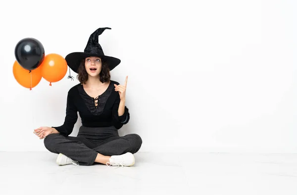 Young witch holding black and orange air balloons sitting on the floor thinking an idea pointing the finger up