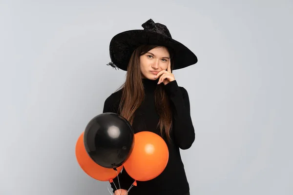 Young witch holding black and orange air balloons thinking an idea
