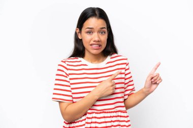Beyaz arka planda izole edilmiş genç Kolombiyalı kadın korkmuş ve kenarı işaret ediyor.