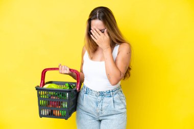 Sarı arka planda, yorgun ve hasta bir ifadeyle izole edilmiş yiyecek dolu bir alışveriş sepeti taşıyan beyaz bir kadın.