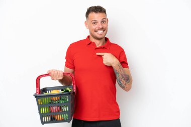 Beyaz arka planda, şaşırtıcı bir yüz ifadesiyle izole edilmiş yiyecek dolu bir alışveriş sepeti tutan beyaz bir adam.