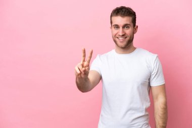 Pembe arka planda izole edilmiş genç beyaz adam gülümsüyor ve zafer işareti gösteriyor.