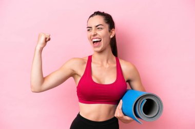 Genç beyaz kadın yoga derslerine gidiyor. Elinde pembe arka planda izole edilmiş bir minder var. Zaferi kutluyor.