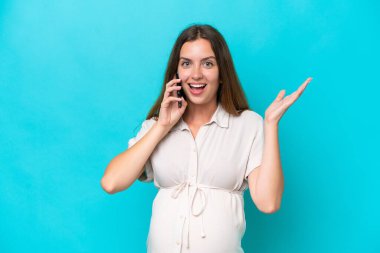 Beyaz bir kadın mavi arka planda izole edilmiş hamile ve şaşırmış bir ifadeyle cep telefonu kullanıyor.