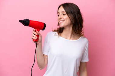 Kafkasyalı genç bir kadın elinde saç kurutma makinesiyle pembe arka planda mutlu bir ifadeyle izole edilmiş.