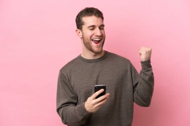 Pembe arka planda izole edilmiş, telefon zafer pozisyonunda genç beyaz bir adam.