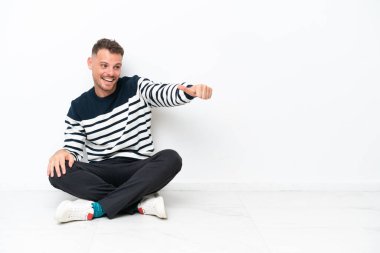 Genç bir adam beyaz arka planda izole bir şekilde yerde oturuyor ve baş parmak işareti yapıyor.