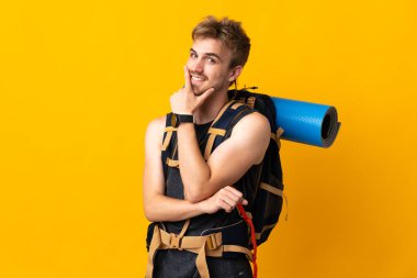 Young mountaineer man with a big backpack isolated on yellow background happy and smiling