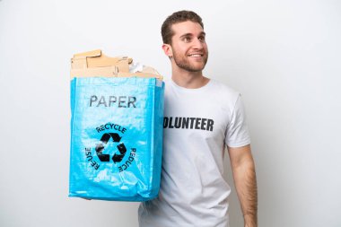 Beyaz arka planda izole edilmiş bir şekilde geri dönüşüm için kağıt dolu bir geri dönüşüm çantası tutan genç beyaz bir adam bir fikir ararken