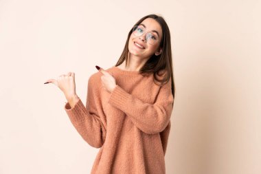 İzole edilmiş bej arka planı olan genç bir kadın bir ürünü sunmak için kenarı işaret ediyor.