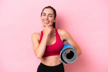 Genç beyaz kadın yoga derslerine gidiyor pembe arka planda izole edilmiş bir minderi tutarken mutlu ve gülümsüyor.