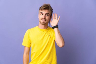Mor arka planda izole edilmiş genç, yakışıklı sarışın bir adam. Kulağına elini sürerek bir şeyler dinliyor.