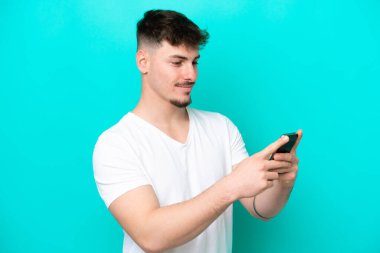 Mavi arka planda izole edilmiş genç beyaz adam cep telefonuyla mesaj ya da e-posta gönderiyor.