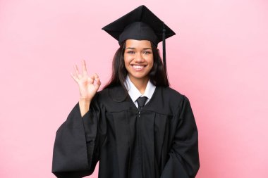Kolombiyalı genç bir üniversite mezunu pembe arka planda izole edilmiş. Parmaklarıyla tamam işareti gösteriyor.