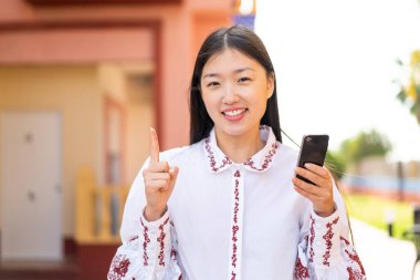 Young Chinese woman using mobile phone at outdoors pointing up a great idea