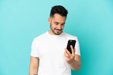 Mavi arka planda izole edilmiş genç beyaz adam cep telefonuyla bir mesaj ya da e-posta gönderiyor