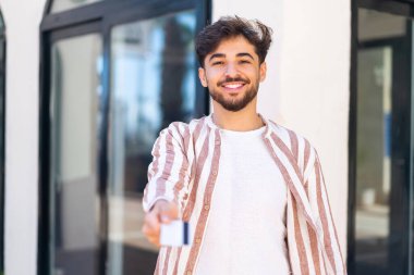 Handsome Arab man at outdoors holding a credit card with happy expression
