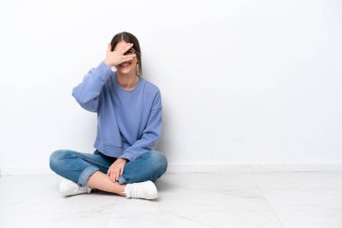 Beyaz arka planda izole bir şekilde yerde oturan genç beyaz kadın gözlerini elleriyle kapatıp gülümsüyor.