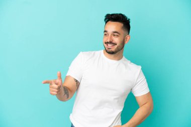 Mavi arka planda izole edilmiş genç beyaz bir adam parmağını yana doğru uzattı ve bir ürün sundu.